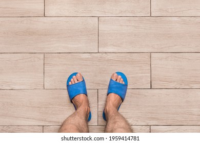 Men's Bare Feet In Blue Home Slippers Stand On A Light Laminate Floor Tile Background, View From Above.