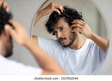 Men's Beauty And Haircare Treatment. Worried Indian Guy Looking At Flaky Scalp In Mirror, Examining Gray Hair And Hairloss Issue, Standing In Bathroom At Home.