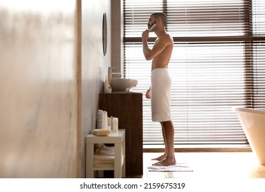 Men's Beauty Routine Concept. Profile Side View Of Handsome Young Man Brushing His Beard With Wooden Comb Looking In The Mirror Standing Wrapped In White Towel In Modern Bathroom, Full Body Length