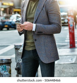 Men`s fashion street style. Classic two buttons jacket green sweatshot and grey jeans. Stylish man walking on city street and texting on cell phone.