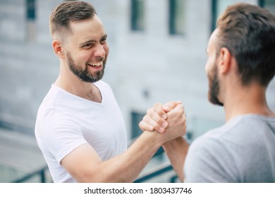 Men's Friendly Handshake. Two Young Bearded Handsome Friends In Casual T-shirts Are Posing Together On The City Background. 
