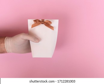 Men's Hand In Medical Glove Holding A White Gift Bag On A Pink Background. Contactless Giving A Present During The Quarantine Period.