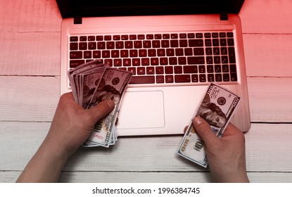 Men's Hands Hold Money Dollars Against The Background Of An Open Laptop. Red Toning. The Concept Of Online Fraud, Internet Fraud, Deception, Extortion Of Money, Debts, Online Gambling.