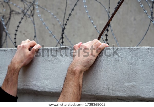 Men\'s hands on a concrete fence against a\
background of barbed wire.  trying to escape from prison. The\
concept of escape from prison, guarding the territory from the\
intruders. illegally\
convicted