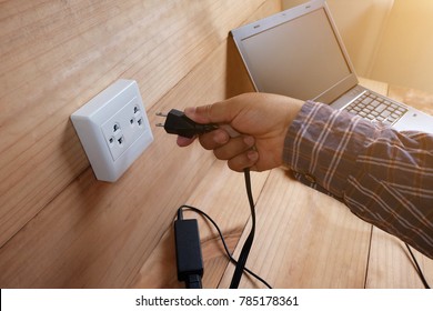 Men's Hands Are Plug In Power Outlet Adapter Cord Charger Of Laptop Computer On Wooden Floor With Sun Warm Light