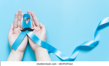 Men's health and Prostate cancer awareness campaign in November. Man hands holding light blue ribbon awareness w/ mustache on blue background. Symbol for support men who living w/ cancer. Copy space.