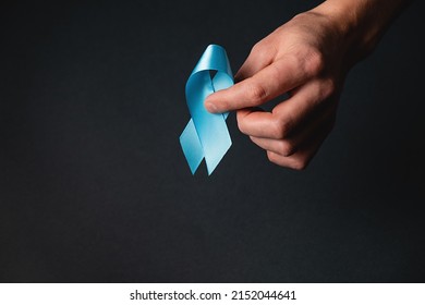 Men's Health And Prostate Cancer Awareness Campaign Concept. Male Hand Holding Blue Ribbon Awareness On Black Background Wall In Studio With Copy Space. Symbol For Support Men Who Living Cancer