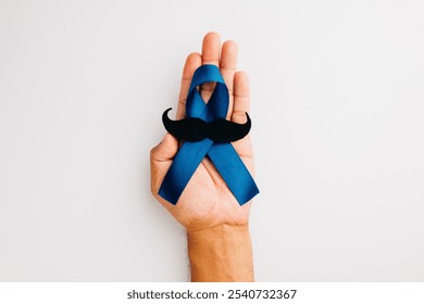 Men's health and Prostate cancer awareness campaign in November. A man's hands clasp a light blue ribbon with a mustache on a blue background, signifying support. - Powered by Shutterstock