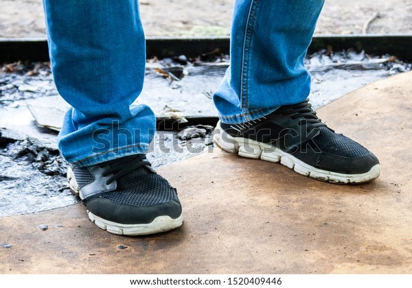 Men\'s legs in blue jeans and black sneakers\
with white soles stand on a dirty oiled plywood board. Dirty car\
master workplace