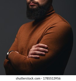 Men's style concept. Cropped portrait of handsome young man with beautiful beard posing in brown turtleneck over dark gray background. Hands crossed. Studio shot