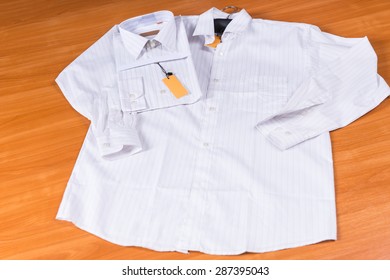 Mens White Dress Shirt Laid Out Flat On Wooden Surface, With Identical Shirt Folded With Tag Attached On Wooden Background