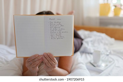 Mental Health Care, Positive Thinking And Wellness Concept. Attractive Woman Lying In Bed, Practicing Stress Relief Challenge With Journal About Feeling, Glass Of Water And Gratitude List