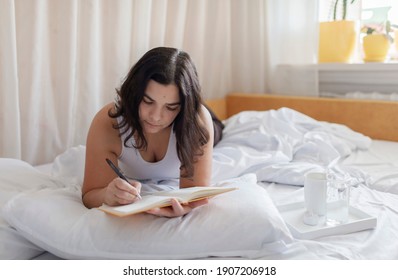 Mental Health Care, Positive Thinking And Wellness Concept. Attractive Woman Lying In Bed, Practicing Stress Relief Challenge With Journal About Feeling, Glass Of Water And Gratitude List