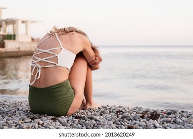 Mental Health Concept. Rear View Of A Depressed Young Woman In Swimsuit Sitting On The Beach, Arms Around Legs