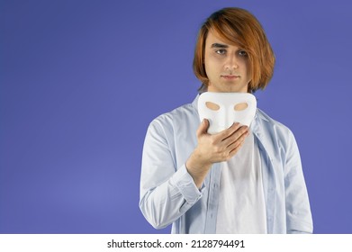 Mental Health Concept. Young Man Holding A Full-face Mask, Ball, Or Masquerade Carnival Mask. Symbol Of Bipolar Disorder Or Hypocrisy, Deceit, Duplicity. Portrait Of Guy Prep Student With Long Hair 