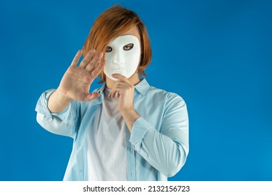 Mental Health Concept. Young Man Holding A Full-face Mask, Ball, Or Masquerade Carnival Mask. Symbol Of Bipolar Disorder Or Hypocrisy, Deceit, Duplicity. Portrait Of Guy Prep Student With Long Hair 