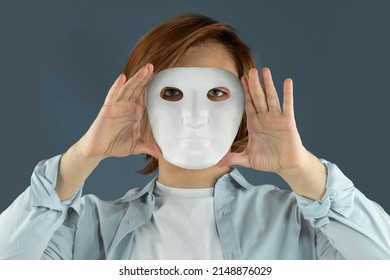 Mental Health Concept. Young Man Holding A Full-face Mask, Ball, Or Masquerade Carnival Mask. Symbol Of Bipolar Disorder Or Hypocrisy, Deceit, Duplicity. Portrait Of Guy Prep Student With Long Hair 