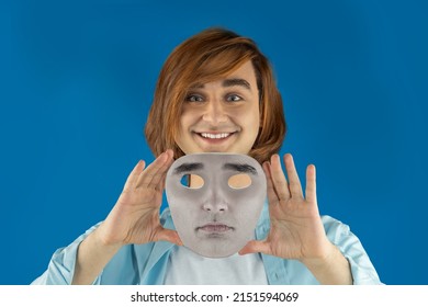 Mental Health Concept. Young Man Holding A Full-face Mask, Ball, Or Masquerade Carnival Mask. Symbol Of Bipolar Disorder Or Hypocrisy, Deceit, Duplicity. Portrait Of Guy Prep Student With Long Hair 