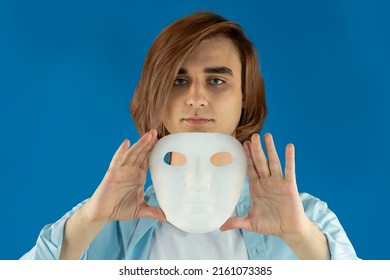 Mental Health Concept. Young Man Holding A Full-face Mask, Ball, Or Masquerade Carnival Mask. Symbol Of Bipolar Disorder Or Hypocrisy, Deceit, Duplicity. Portrait Of Guy Prep Student With Long Hair 