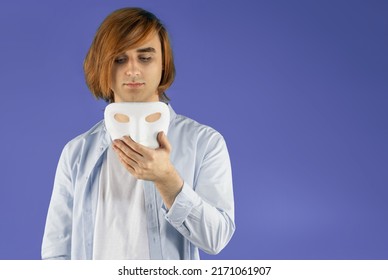 Mental Health Concept. Young Man Holding A Full-face Mask, Ball, Or Masquerade Carnival Mask. Symbol Of Bipolar Disorder Or Hypocrisy, Deceit, Duplicity. Portrait Of Guy Prep Student With Long Hair 