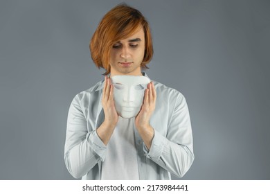 Mental Health Concept. Young Man Holding A Full-face Mask, Ball, Or Masquerade Carnival Mask. Symbol Of Bipolar Disorder Or Hypocrisy, Deceit, Duplicity. Portrait Of Guy Prep Student With Long Hair 