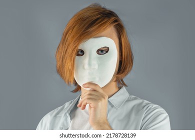 Mental Health Concept. Young Man Holding A Full-face Mask, Ball, Or Masquerade Carnival Mask. Symbol Of Bipolar Disorder Or Hypocrisy, Deceit, Duplicity. Portrait Of Guy Prep Student With Long Hair 