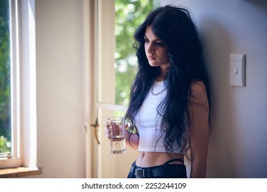 Mental health, depression and sad woman thinking with water, stress and anxiety in a house. Depressed, lonely and goth girl with a drink, emotional and comtemplating problems in the lounge of home