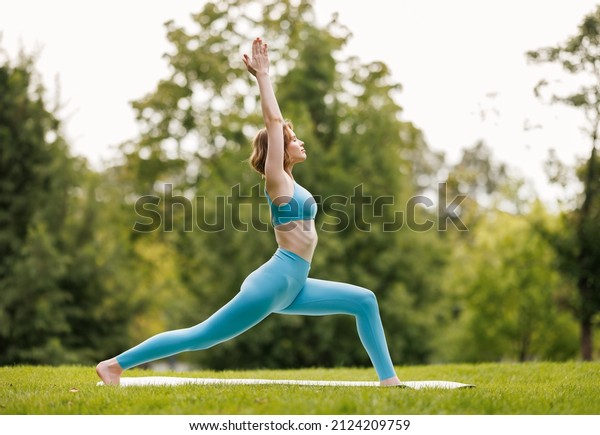 Mental and\
physical health. Slender focused girl in sportswear performing yoga\
outside in early morning in nature, female standing in warrior  \
pose with hands raised on grass in city\
park