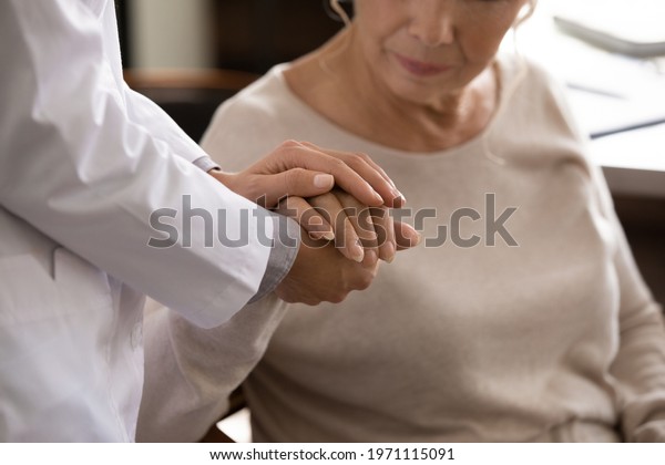 Mental support. Close up of disabled sick senior\
female hand in palms of young woman in white medical uniform.\
Attending physician nurse comforting taking care of unhealthy aged\
lady geriatric patient