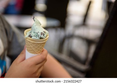 Menthol Ice Cream With Chocolate Chips Close-up. A Child's Hand Holds A Delicious Ice Cream Cone On The Background Of A Summer Cafe.