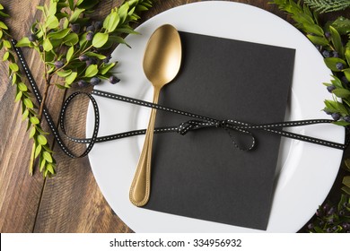 menu place setting with empty card and golden spoon over wooden background, surrounded by green branches