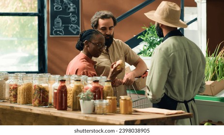 Merchant assists customers in grocery store, showing freshly harvested fruits and vegetables. Young couple searching for organic produce at local zero waste supermarket, sustainable lifestyle. - Powered by Shutterstock