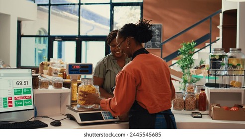 Merchant weighting bulk items on scale for regular client, selling various types on homemade products in reusable jars at checkout. Vendor chatting with buyer about healthy eating. Handheld shot. - Powered by Shutterstock