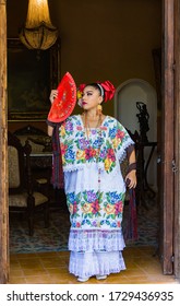 Merida,Yucatan/Mexico-February 29,2020: Woman In Traditional Embroidered Huipil Tunic And Dress Stands With Her Red Fan In Hacienda's Doorway 
