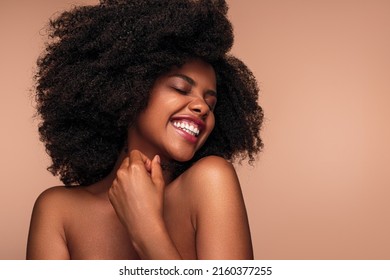 Merry African American woman with bare shoulders and Afro hairstyle smiling with closed eyes and touching neck after spa procedure against brown background - Shutterstock ID 2160377255