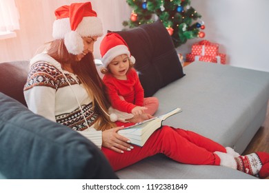 Merry Christamas And Happy New Year. Young Woman With Red Hat Sit On Sofa With Daughter. She Holds Book On Her Lap. Woman Looks Down And Smiles. Small Girl Is Calm. She Looks At Book Too.