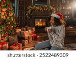 Merry Christmas. African American woman in Santa hat with cup of hot drink coffee tea near Christmas tree in dark interior. Girl in living room with Christmas tree and fireplace. Christmas eve at home