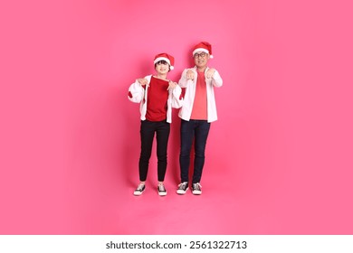 Merry Christmas, Asian senior and young woman wear santa hat and casual red clothes with white heart-shaped shaped shirt jacket with gesture of pointing, isolated on the pink background. - Powered by Shutterstock