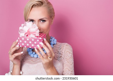 Merry Christmas. Beautiful Blonde Woman Holding Small Gift Box With Ribbon. Soft Colors. Studio Portrait Over Pink Background