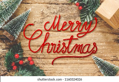 Merry Christmas Card On Wood Floor Top View Handwriting Red Xmas On Wooden Table With Mini Christmas Tree And Mistletoe And Home Toy Decoration With Snow Fall.happy Winter Holiday Festive Seasonal