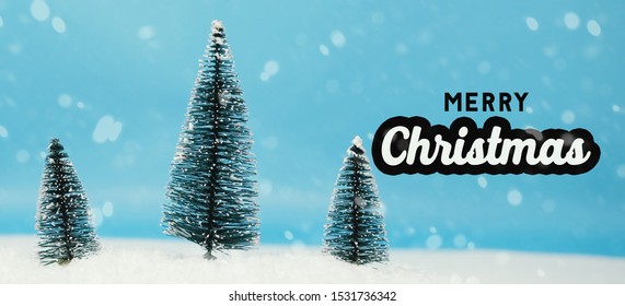 Merry Christmas Celebration With Bottle Brush Christmas Tree Decoration In Snow, Text On Blue Background.