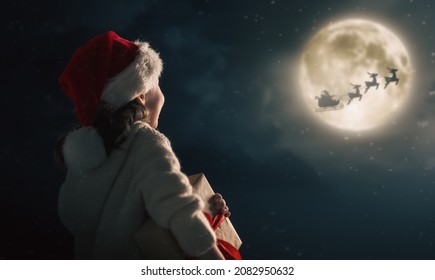 Merry Christmas. Cute little child with present. Santa Claus flying in his sleigh against moon sky. Happy kid enjoy the holiday.  - Powered by Shutterstock