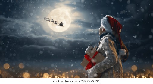 Merry Christmas. Cute little child with xmas present. Santa Claus flying in his sleigh against moon sky. Happy kid enjoy the holiday. Portrait of girl with gifts on dark background.