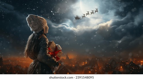 Merry Christmas. Cute little child with xmas present. Santa Claus flying in his sleigh against moon sky. Happy kid enjoy the holiday. Portrait of girl with gifts on dark background. - Powered by Shutterstock