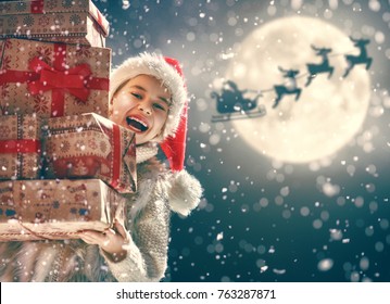 Merry Christmas! Cute little child with xmas present. Santa Claus flying in his sleigh against moon sky. Happy kid enjoy the holiday. Portrait of girl with gifts on dark background.