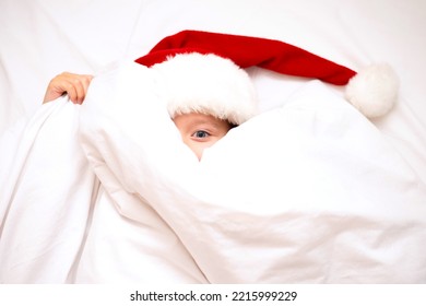 Merry Christmas. A funny girl in Santa's hat peeks out from under the covers in bed. White background. Emotions. Merriment. Hotel. - Shutterstock ID 2215999229