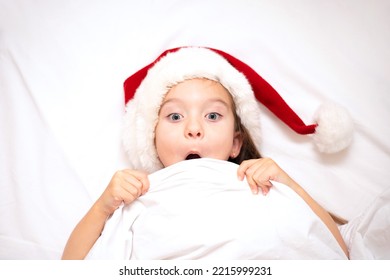 Merry Christmas. A funny girl in Santa's hat peeks out from under the covers in bed. White background. Emotions. Merriment. Hotel. - Shutterstock ID 2215999231