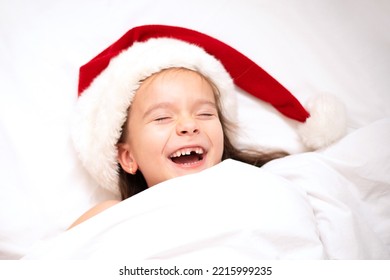 Merry Christmas. A funny girl in Santa's hat peeks out from under the covers in bed. White background. Emotions. Merriment. Hotel. - Shutterstock ID 2215999235