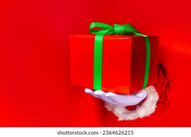 Merry Christmas greeting card bright red background, gifts and sale promotion banner background. Santa hands holds  big red gift box with festive green ribbon in red torn paper hole background - Shutterstock ID 2364626215
