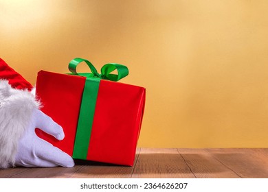 Merry Christmas greeting card, gifts and sale promotion banner background. Santa hands with big red gift box with festive green ribbon, over gold background - Shutterstock ID 2364626207